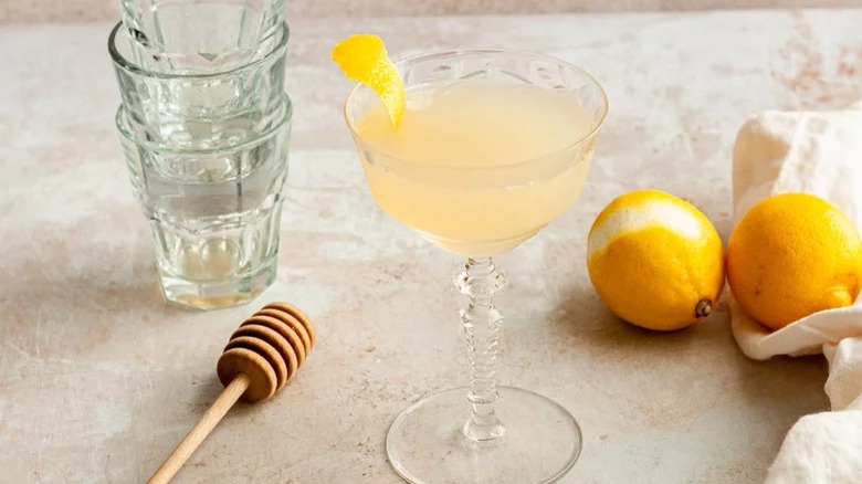 Bee's Knees cocktail in a coupe glass, garnished with lemon peel, with lemons and a honey dipper in the background