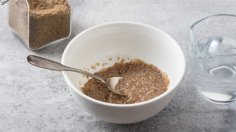 A white bowl of ground flax and water that has begun congealing like a raw egg, next to a glass jar of flaxseed and a glass water pitcher