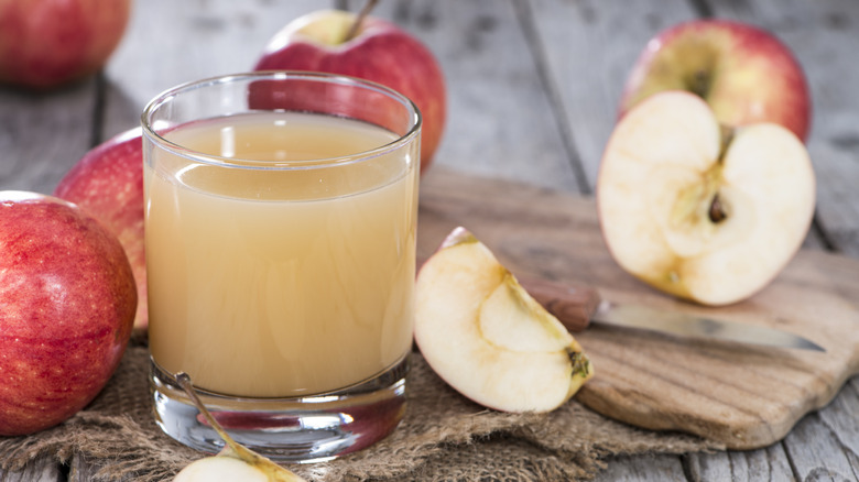 A short glass of apple juice, surrounded by whole and cut apples, with a cutting board and knife in the background