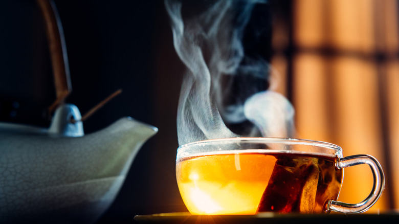 A clear mug filled with steaming hot, golden-colored tea, with sunlight shining through it and a blue teapot to the left