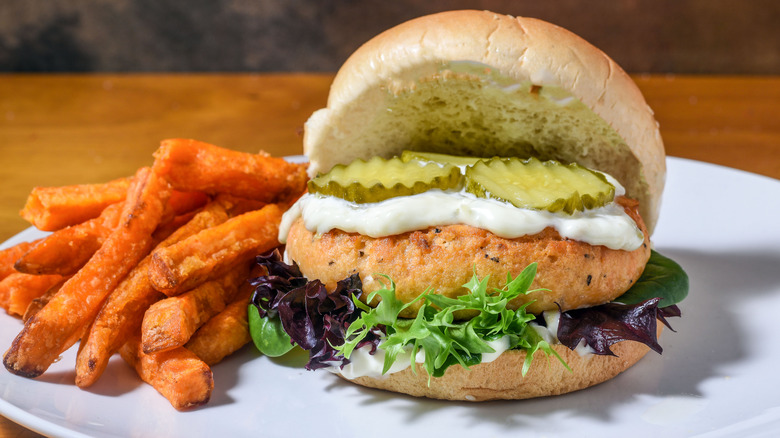 Salmon burger with lettuce, tartar sauce, and pickles on a plate with sweet potato fries