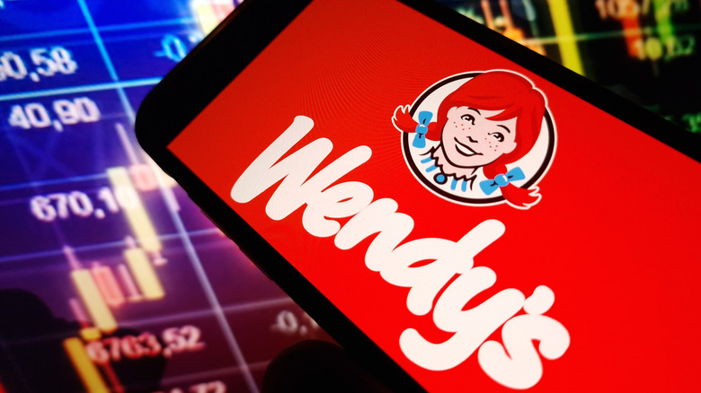 A smartphone with the Wendy's logo on it, in front of a background of stock ticker information.