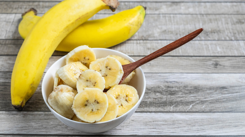 Bowl of banana slices with two bananas in background.