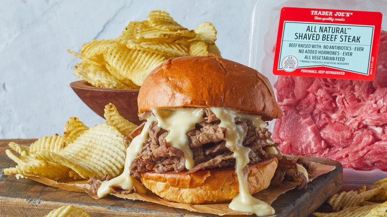 a steak sandwich on thick brioche bun with molten cheese running down the sides and a bowl of potato chips sits in front of a package of Trader Joe's All Natural Shaved Beef Steak.