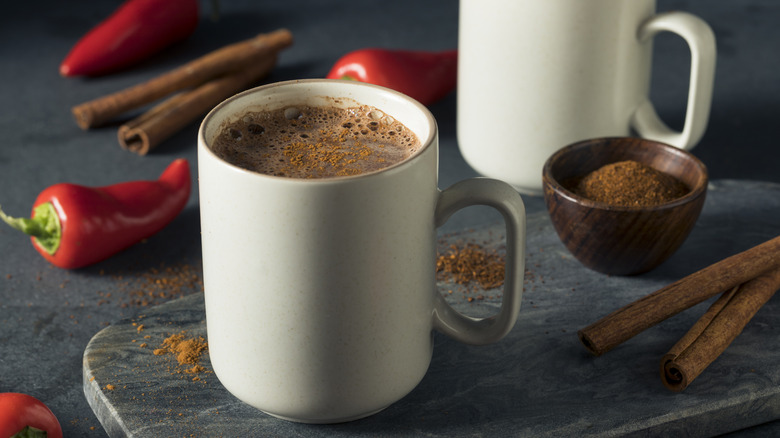 Cup of hot chocolate topped with spices, surrounded by cayenne peppers and cinnamon sticks.