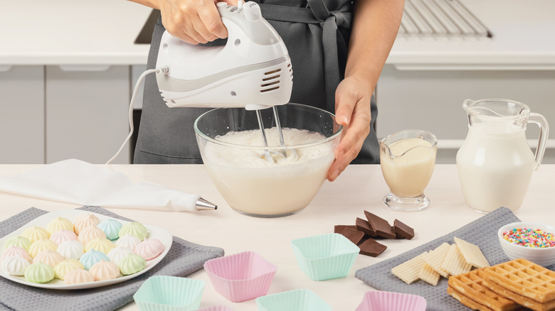 A person mixing together an ice cream base with a hand mixer, and mix-ins spread out on a table