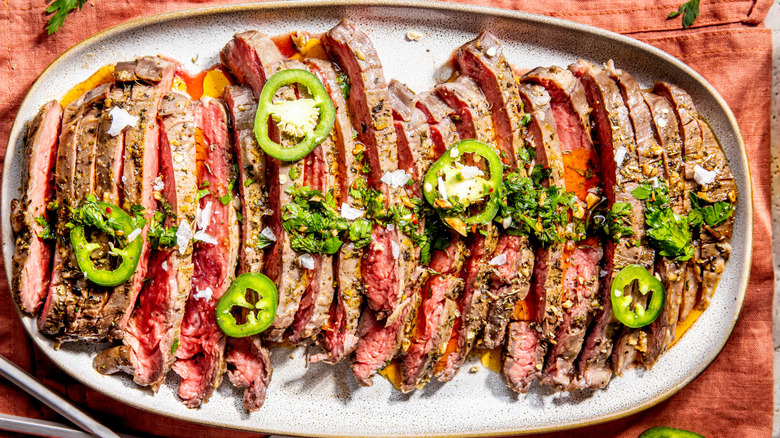 Grilled, marinated, sliced flank steak on an oval plate, garnished with jalapeño slices and fresh herbs