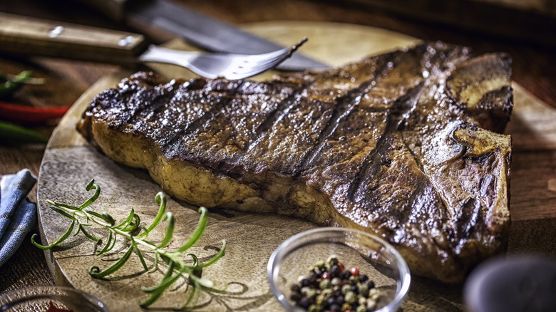 Grilled t-bone steak on a wooden cutting board, with rosemary and peppercorns