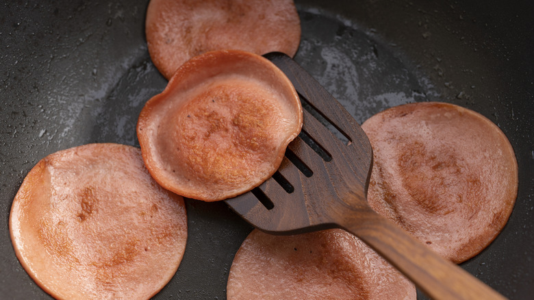 closeup of bologna frying in a pan, with a wooden spatula lifting one slice