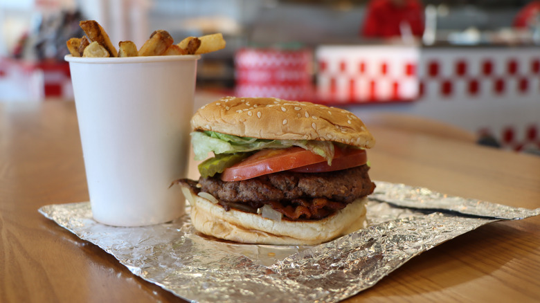 a Five Guys burger with lettuce, pickles, tomato, bacon, and onions on a table with a cup of fries.