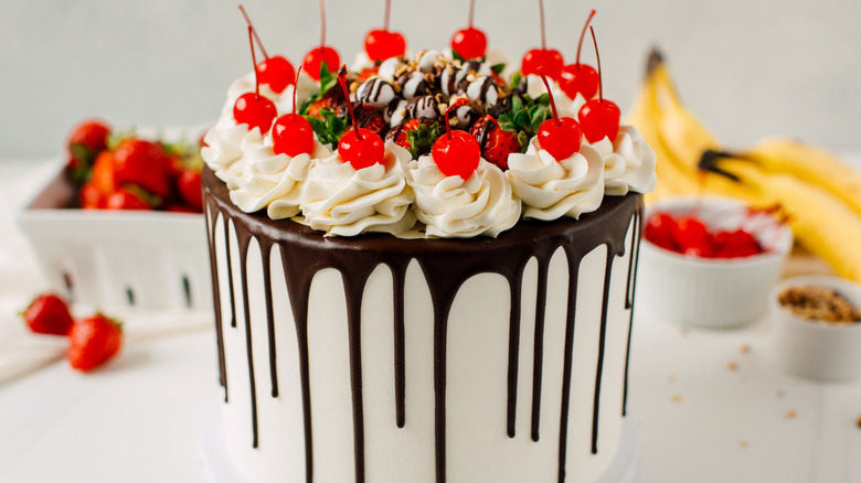 A banana split cake with chocolate syrup dripping down the sides topped with cherries and dollops of whipped cream.