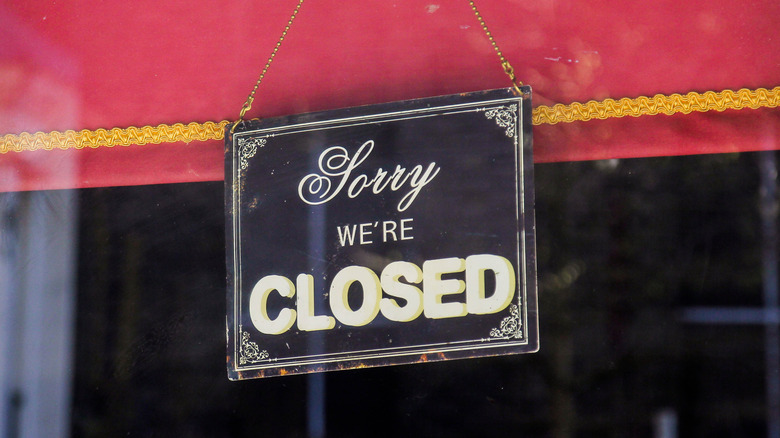 A "sorry, we're closed" sign in front of a red curtain at a dark, closed store