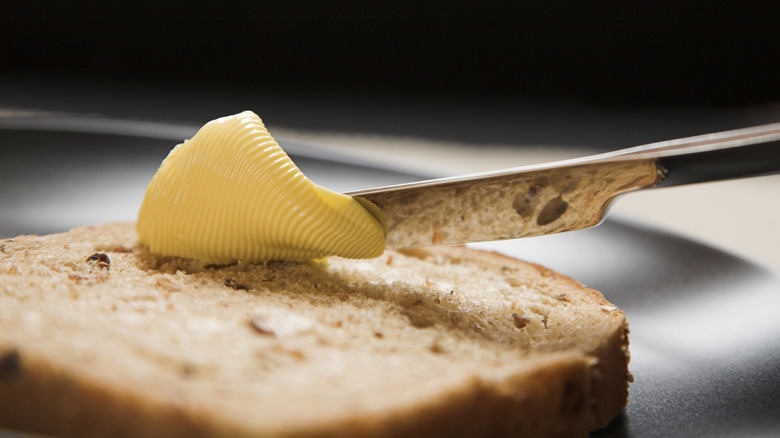 A yellow square of spreadable butter being applied by silver butter knife to a brown slice of bread