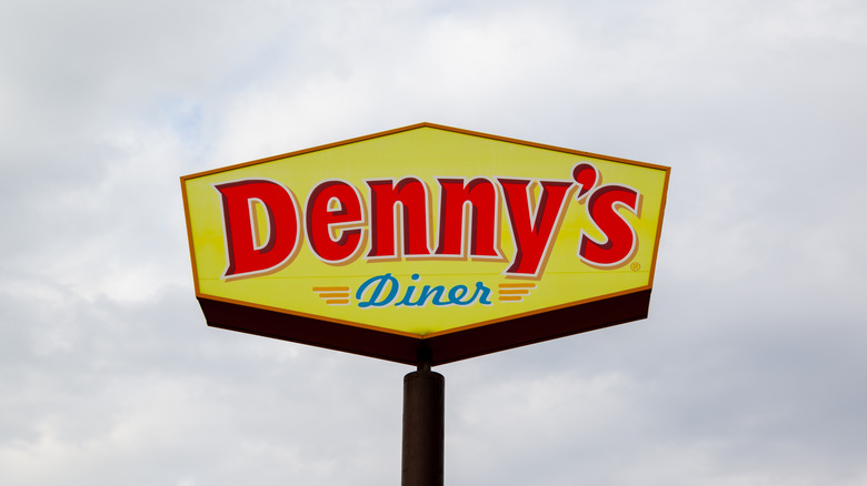 A standalone sign that says "Denny's Diner" in red and blue text, on a yellow background, against a cloudy gray sky