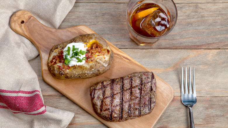 Steak and a baked potato sitting on a cutting board, with a glass of bourbon next to it