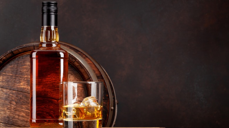Bottle of whiskey in front of barrel, with glass of whiskey on ice