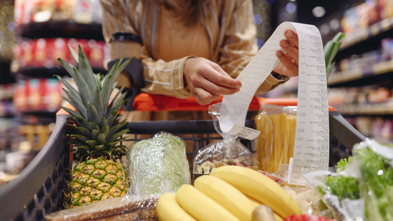A shopper reviews their purchase receipt at a grocery store while pushing a cart full of fresh fruit