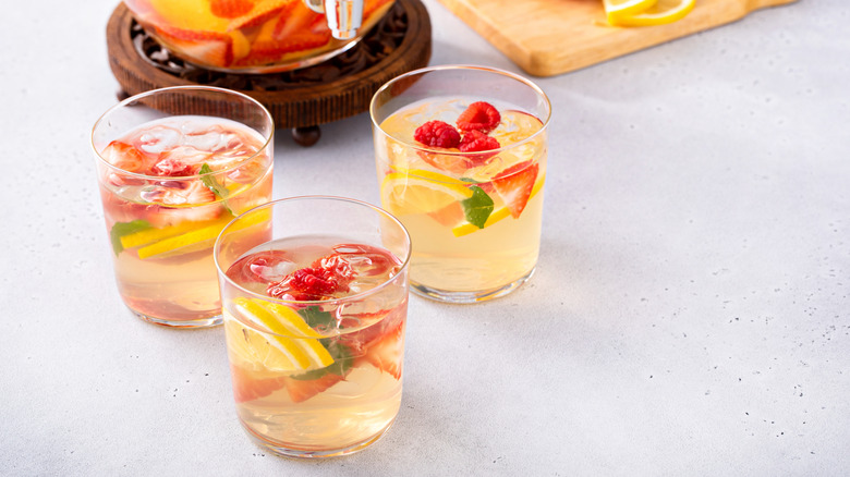 Three glasses of white wine sangria with lemonade pitcher and fruit on cutting board