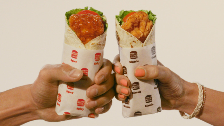 Two outstretched hands holding Classic and Spicy Royal Crispy wraps from Burger King, toasting each other with the wraps.