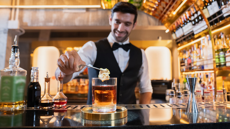 A bartender smiles while adding garnish to a cocktail at a well-stocked bar