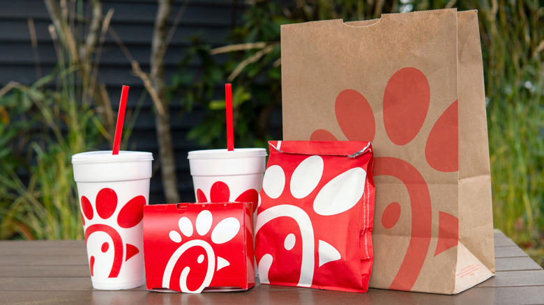 Chick-fil-A bag, drinks, bag, and box on picnic table with greenery and wall in background