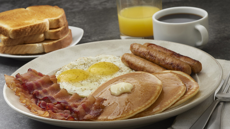 A classic American breakfast plate with pancakes, bacon, fried eggs, and sausages, with toast, juice, and coffee on the side