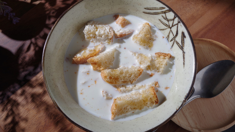 A bowl with milk toast on a wooden table with a spoon on the side