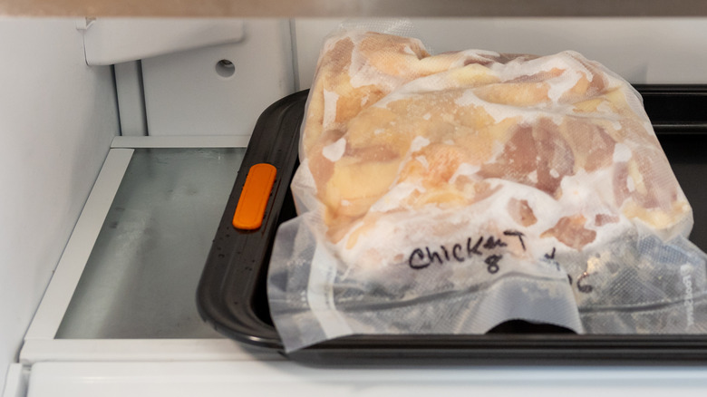 A bag of frozen chicken pieces thawing on a black cookie sheet in a refrigerator