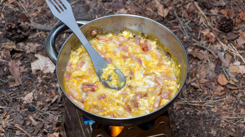 Scrambled eggs and bacon cooking in a pan over an open fire
