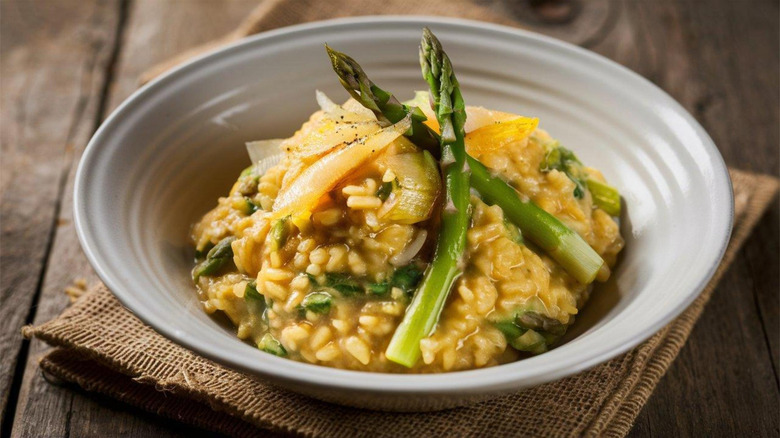 A white bowl of risotto with veggies on top, resting on a wooden table