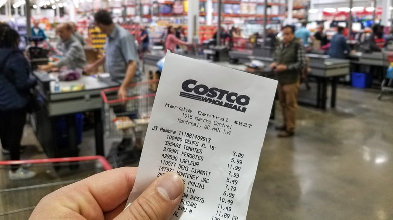 Hand holding Costco receipt with store registers and customers blurred in the background