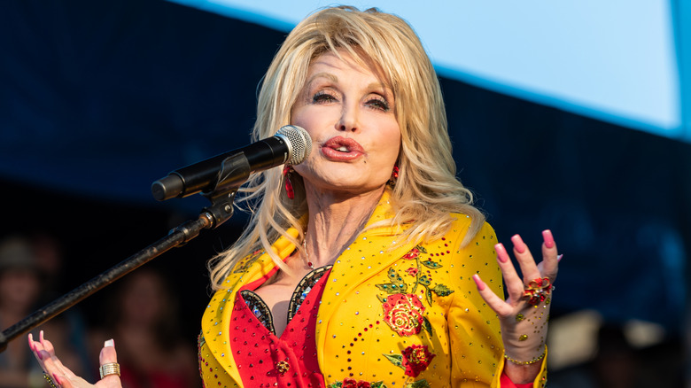 Dolly Parton in a sequined yellow jacket, singing into a standing microphone