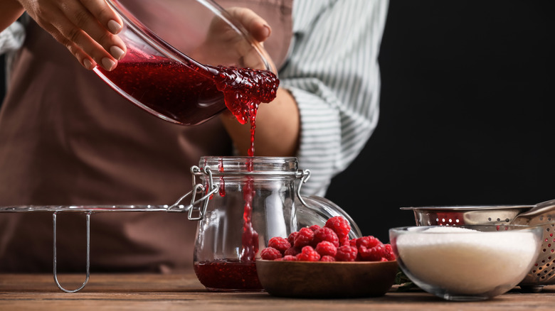 Chef pouring cooled jam from glass bowl into a glass jar with bowls of fresh raspberries and sugar