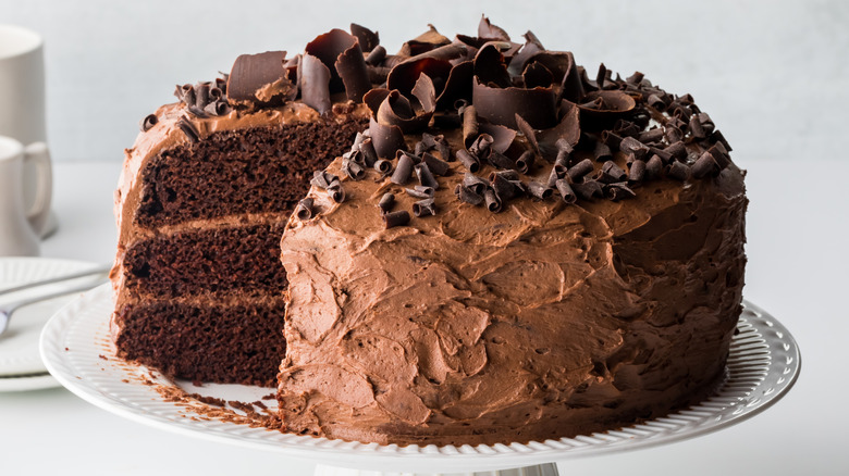 Chocolate cake with a missing slice on white cake stand with white dishes in the background