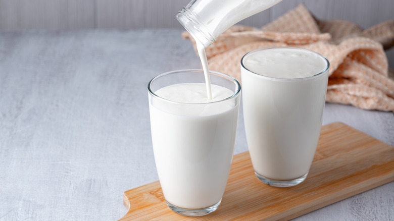 Pouring homemade buttermilk (sour milk) into glasses on a narrow serving board with a blurred towel in the background
