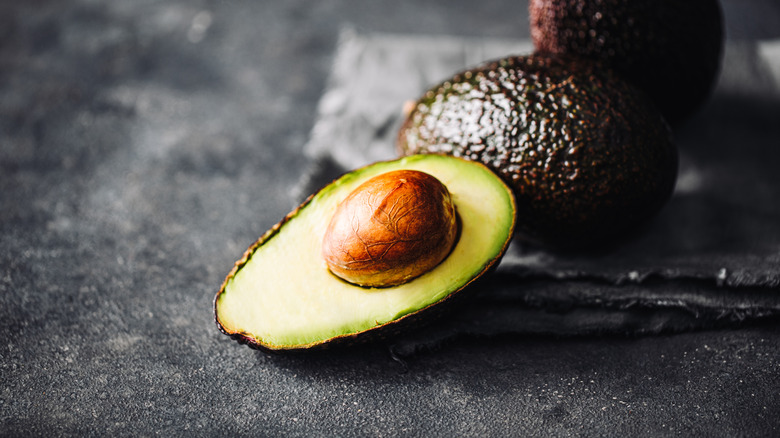 Half an avocado on a slate background with two whole avocados resting behind it