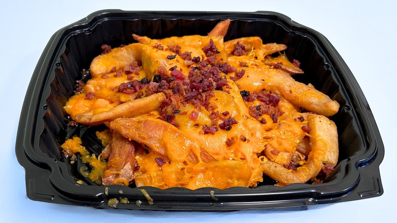 Cheese fries topped with cheddar and bacon in a black container against a light blue background