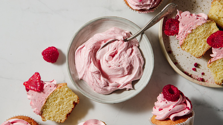 Bowl of pink frosting with spoon in it surrounded by raspberry cupcakes
