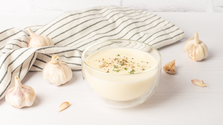 Aioli in transparent bowl, alongside garlics on a white table