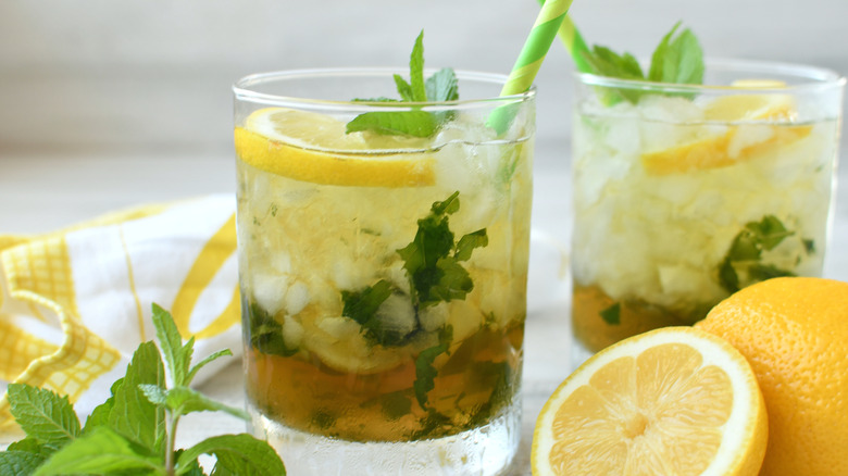 Glass of water with muddled mint and lemon with a green straw surrounded by lemon slices and mint.