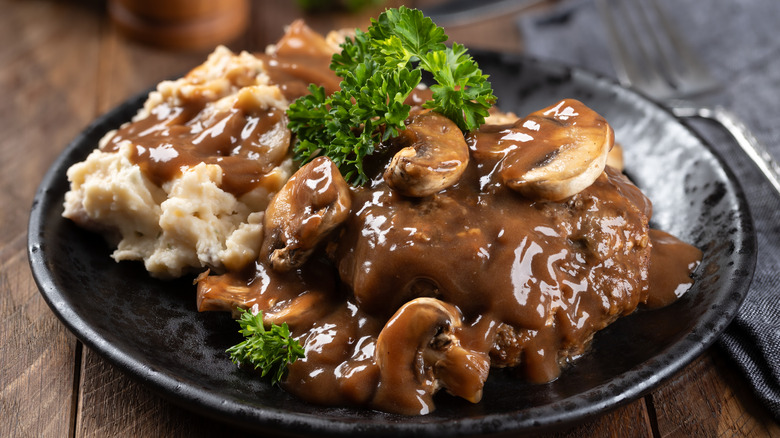 Old-school Salisbury steak topped with mushroom gravy and a side of mashed potatoes on a black plate