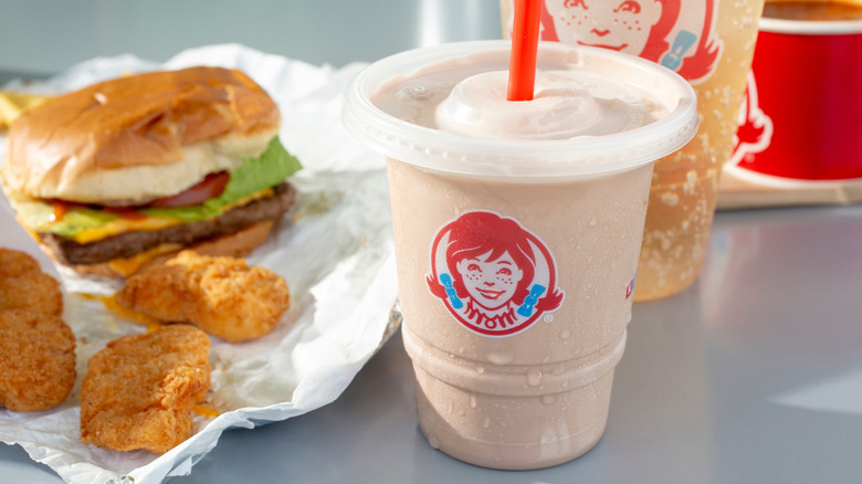 Wendy's chocolate Frosty with a burger and chicken nuggets on a table with a soda in the background