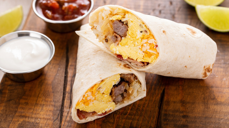 Breakfast burrito cut in half on a wood board with salsa and sour cream on side