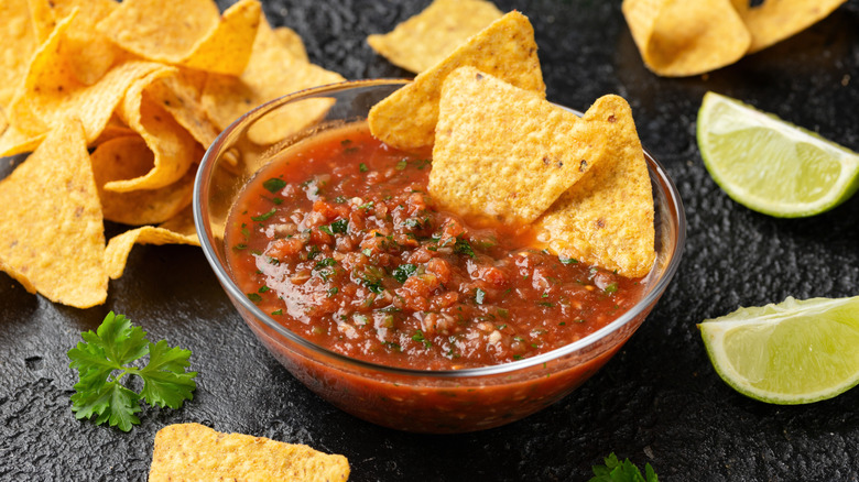 A bowl of salsa with corn chips, next to cilantro and limes