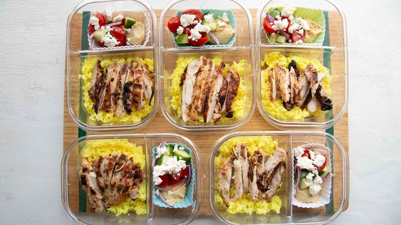 five meal prep containers with chicken, rice, and silicone muffin tin with hummus and tomato cucumber feta salad