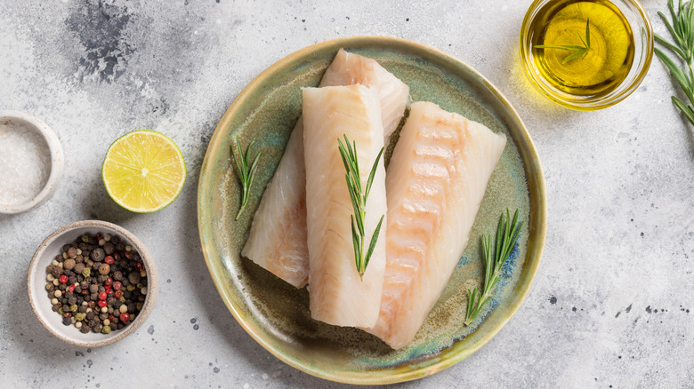 A ceramic plate with three raw cod filets topped with rosemary sprigs and lemon olive oil and peppercorns on the side.