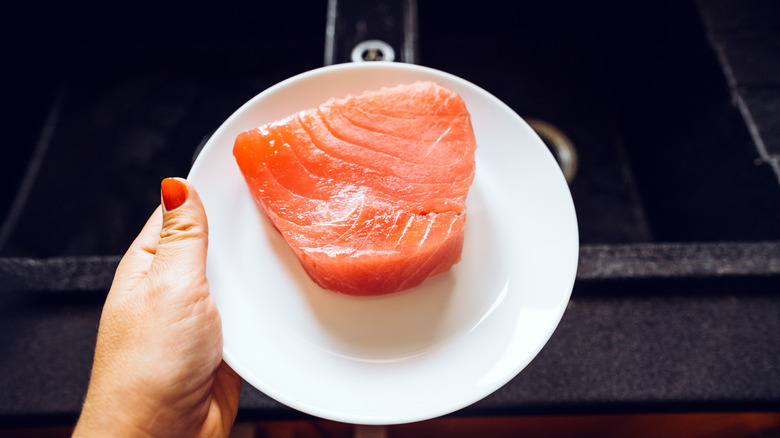 A hand holding a white plate with a raw tuna steak.