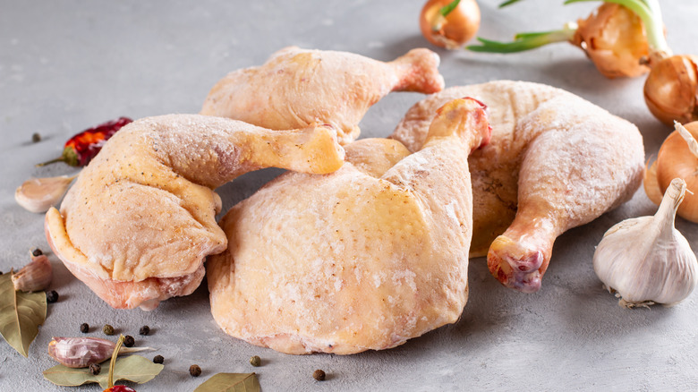 Frozen chicken legs surrounded by garlic, peppers, onions, peppercorns, and bay leaves on gray counter