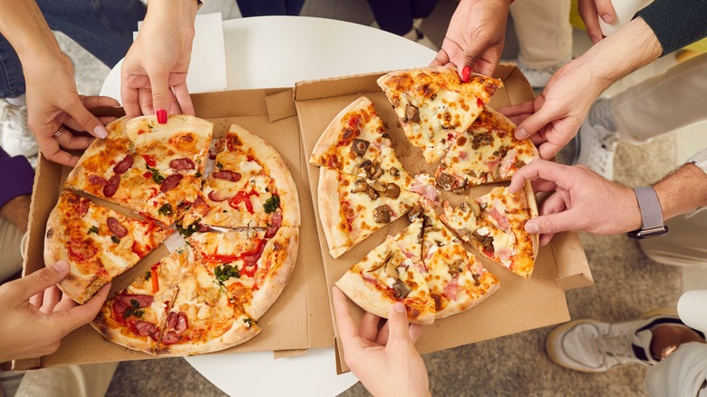 An overhead view of two pizzas with several pairs of hands grabbing a slice each