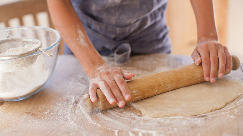 A woman rolling out dough on a wooden surface with flour dusted on it and a bowl of flour on the side